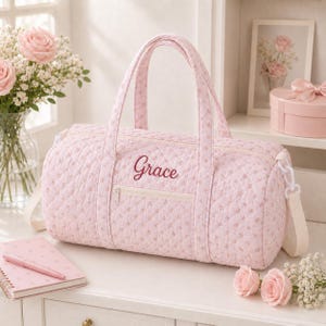 Puede incluir: Un bolso de lona acolchado de color rosa claro con un pequeño estampado floral y el nombre Grace bordado en letra cursiva rosa oscuro en el bolsillo frontal con cremallera. Incluye dos asas superiores y una correa blanca desmontable.