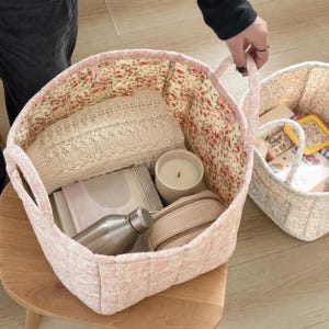 May include: A quilted fabric storage basket with a pink and white floral exterior and a red and green floral interior lining. The basket contains a beige knit sweater, a white candle, a metallic water bottle, and a beige zippered pouch.
