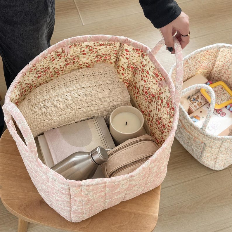 May include: A quilted fabric storage basket with a pink and white floral exterior and a red and green floral interior lining. The basket contains a beige knit sweater, a white candle, a metallic water bottle, and a beige zippered pouch.