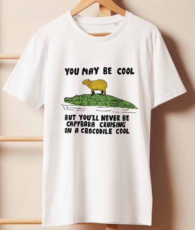 May include: White t-shirt with a graphic of a capybara riding a crocodile. The text on the shirt reads "You may be cool but you'll never be capybara cruising on a crocodile cool."