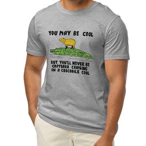 May include: A gray t-shirt with the text "You may be cool but you'll never be capybara cruising on a crocodile cool."  A cartoon image of a capybara riding a crocodile is above the text.