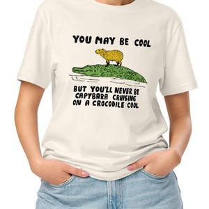 May include: A white t-shirt with a black graphic of a capybara riding a crocodile. The text on the shirt reads "You may be cool but you'll never be capybara cruising on a crocodile cool."