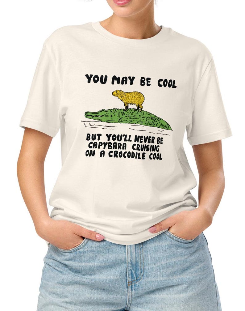 May include: A white t-shirt with a black graphic of a capybara riding a crocodile. The text on the shirt reads "You may be cool but you'll never be capybara cruising on a crocodile cool."