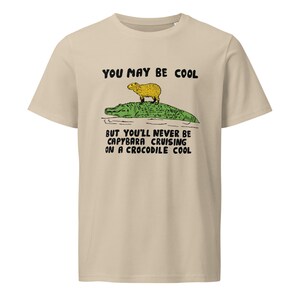 May include: Beige t-shirt with a graphic of a capybara riding a crocodile. The text on the shirt reads "You may be cool but you'll never be capybara cruising on a crocodile cool."