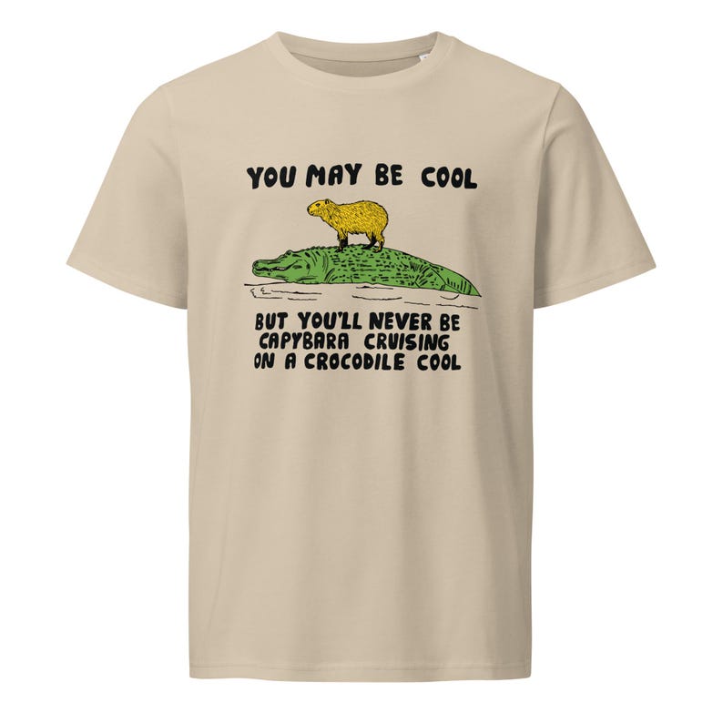 May include: Beige t-shirt with a graphic of a capybara riding a crocodile. The text on the shirt reads "You may be cool but you'll never be capybara cruising on a crocodile cool."