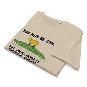 May include: Beige t-shirt with a graphic of a capybara riding a crocodile. The text on the shirt reads "You may be cool but you'll never be capybara cruising."