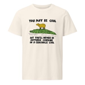 May include: A white t-shirt with a green alligator and a yellow capybara on top. The text "You may be cool but you'll never be capybara cruising on a crocodile cool" is printed on the shirt.