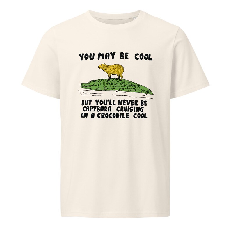 May include: A white t-shirt with a green alligator and a yellow capybara on top. The text "You may be cool but you'll never be capybara cruising on a crocodile cool" is printed on the shirt.