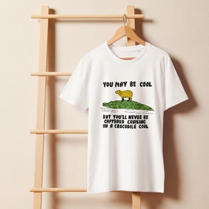 May include: White t-shirt with a graphic of a capybara riding a crocodile. The text on the shirt reads "You may be cool but you'll never be capybara cruising on a crocodile cool."