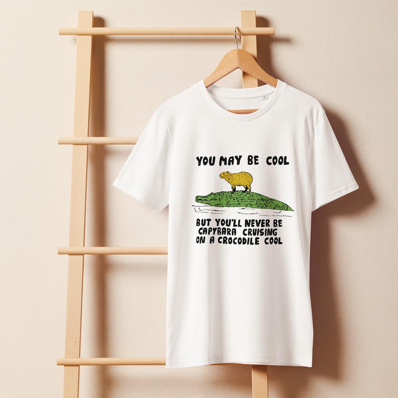 May include: White t-shirt with a graphic of a capybara riding a crocodile. The text on the shirt reads "You may be cool but you'll never be capybara cruising on a crocodile cool."