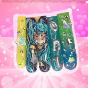 May include: A set of five long, decorated artificial nails. The nails feature designs including a cartoon character, musical notes, and a star. The nails are painted in shades of yellow, blue, and green, with silver accents and the text "FATAL FEMME NAILS" at the top.