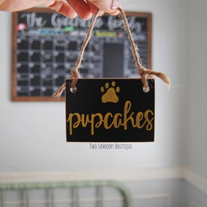 May include: A black wooden sign with gold lettering that reads "pupcakes" and a gold paw print. The sign is hanging from a twine string and has the text "Two Shmoops Boutique" at the bottom.