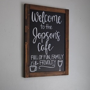May include: A brown wooden framed chalkboard sign with white lettering that reads "Welcome to the Jopson's Cafe Full of Fun, Family & Frivolity". The sign has a coffee cup and wine glass graphic on the bottom.