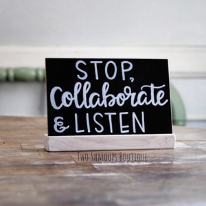May include: Black chalkboard sign with white text that reads "STOP, Collaborate & LISTEN". The sign is on a wooden stand and has the words "TWO SHMOOPS BOUTIQUE" written below the sign.