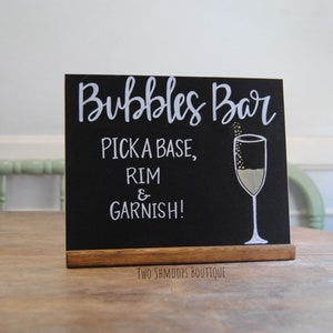 May include: A black chalkboard sign with white lettering that reads "Bubbles Bar, Pick a Base, Rim & Garnish!" A champagne glass illustration is on the right. The sign is on a wooden base.