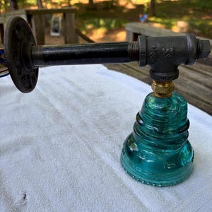 May include: A wall-mounted light fixture featuring a black industrial pipe arm attached to a circular flange. The fixture includes a T-shaped pipe fitting connected to a brass-colored collar, which supports a translucent teal glass insulator.