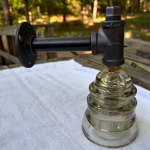 May include: A wall-mounted light fixture featuring a black industrial pipe arm with a circular mounting flange and a threaded T-joint connector. A clear, tiered glass electrical insulator is attached to the bottom of the pipe assembly.