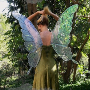 May include: A pair of iridescent butterfly wings with a green and blue colour scheme. The wings are made of a sheer, translucent material and have a delicate, intricate design. The wings are attached to a person's back, creating a whimsical and magical look.