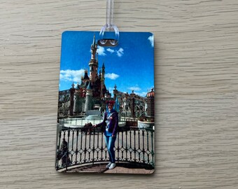 May include: A luggage tag featuring a photograph of a person standing in front of a castle. The castle is light blue with white detailing and a tall, pointed spire. The person is wearing a blue shirt and jeans. The sky is blue with white clouds.