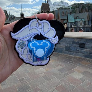 May include: A black Mickey Mouse-shaped keyring with a blue and white design. The keyring features a blue dummy with a white button. The design includes white embroidery and a silver ring.