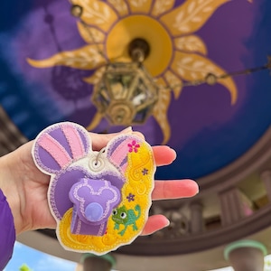 May include: A Disney-themed keychain in purple and yellow, featuring a Mickey Mouse head design with Rapunzel's hair and Pascal the chameleon. The keychain has a button and a small loop for attaching to keys or a bag. The background is a blurred image of a decorative ceiling.