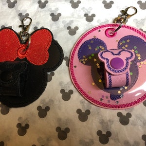 May include: Two keychains featuring a Mickey Mouse design. One keychain is black with a red bow and glitter. The other keychain is pink with purple ears and glitter. Both keychains have a clear plastic pocket for a hand sanitizer.