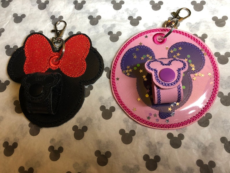 May include: Two keychains featuring a Mickey Mouse design. One keychain is black with a red bow and glitter. The other keychain is pink with purple ears and glitter. Both keychains have a clear plastic pocket for a hand sanitizer.