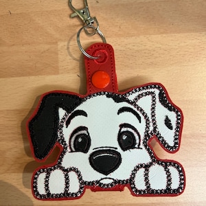Peut inclure: Un chien dalmatien blanc et noir avec des coutures rouges autour des bords, qui sort d'une boucle en tissu rouge avec un porte-clés en métal argenté. Le chien a des yeux noirs et un nez noir.