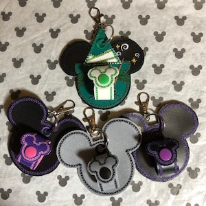 May include: Four different Mickey Mouse-shaped keychains with a small pocket on the side. One keychain is green and features a wizard hat, another is black with a pink pocket, another is gray with a black pocket, and the last is black with a purple pocket.