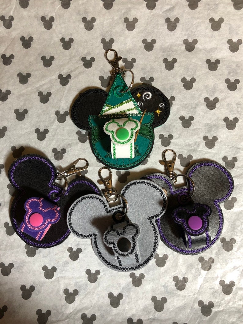 May include: Four different Mickey Mouse-shaped keychains with a small pocket on the side. One keychain is green and features a wizard hat, another is black with a pink pocket, another is gray with a black pocket, and the last is black with a purple pocket.