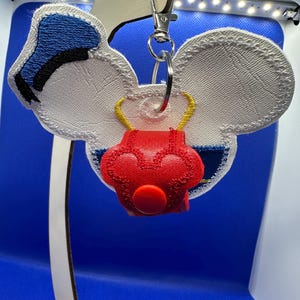 May include: A white and blue embroidered keychain with a red heart-shaped charm. The keychain is shaped like a cartoon character with large ears and a blue hat.