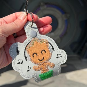 May include: A grey keychain featuring a cartoon character with orange skin, green shorts, and headphones. The character is surrounded by musical notes. The keychain has a silver clasp.