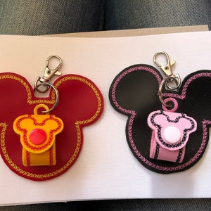 May include: Two Mickey Mouse shaped keychains with a small pouch attached to each. One keychain is red with yellow and red trim and a red button. The other keychain is black with pink and black trim and a white button.