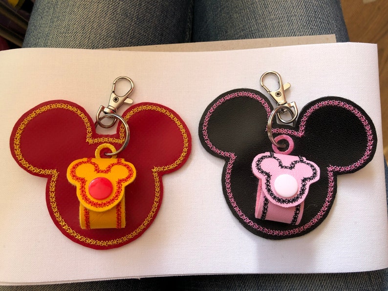 May include: Two Mickey Mouse shaped keychains with a small pouch attached to each. One keychain is red with yellow and red trim and a red button. The other keychain is black with pink and black trim and a white button.