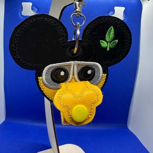 May include: A black and yellow Mickey Mouse-shaped keyring with a green leaf design. The keyring has a silver ring and a yellow button.