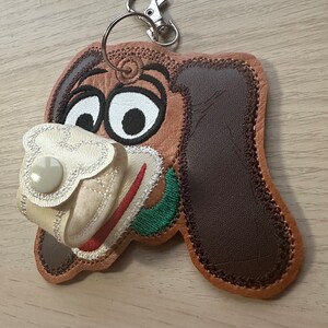 May include: A brown and white embroidered dog keychain with a white fabric pocket and a button. The keychain has a silver metal ring and clip.