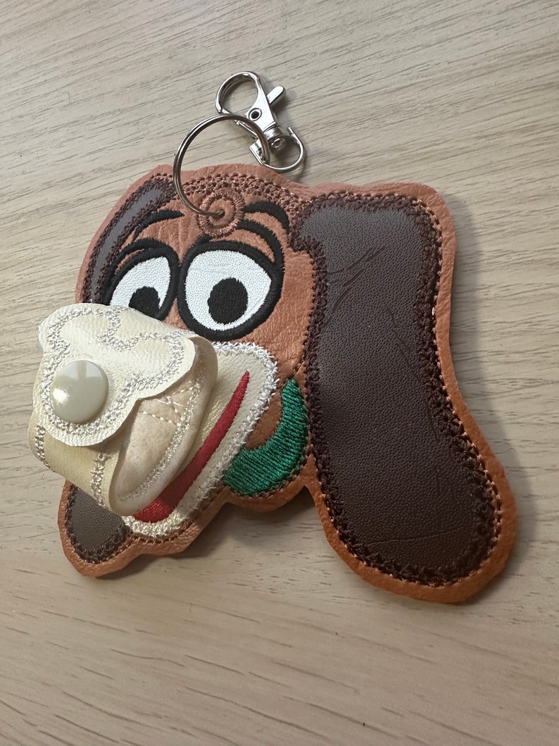 May include: A brown and white embroidered dog keychain with a white fabric pocket and a button. The keychain has a silver metal ring and clip.