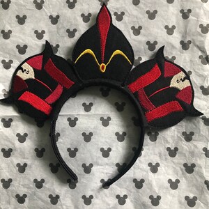 Puede incluir: Diadema negra con orejas de tela roja y dorada. Las orejas tienen forma de corona con una punta puntiaguda y un borde dorado.