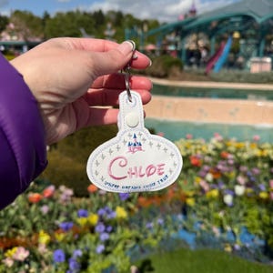 May include: White leatherette keyring with the name "Chloe" embroidered in pink script. The keyring features a castle design and the phrase "WHERE DREAMS COME TRUE". It is attached to a silver-coloured metal clasp.