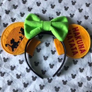 May include: A pair of yellow felt mouse ears with black embroidery of a lion and the words "Hakuna Matata". The ears are attached to a black headband with a green bow.