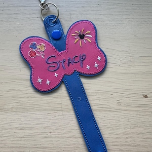 May include: A pink and blue keychain with the name "Stacy" embroidered on it. The keychain features a bow-shaped design with embroidered fireworks and balloons. The strap is blue with a snap closure and a silver clasp.