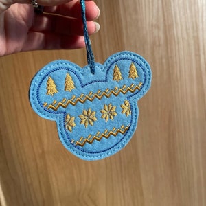 May include: Blue felt Christmas ornament shaped like Mickey Mouse ears with gold embroidered snowflakes and pine trees.