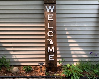 May include: A wooden welcome sign with the word "WELCOME" in white capital letters. The "O" is replaced with a white outline of the state of Michigan. The sign is made of dark brown wood and is placed outdoors.