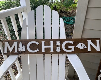 Puede incluir: Un letrero de madera con la palabra "MICHIGAN" en letras blancas. El letrero presenta iconos blancos de árboles, linternas y tiendas de campaña. El letrero es marrón con letras blancas y descansa sobre una silla blanca.