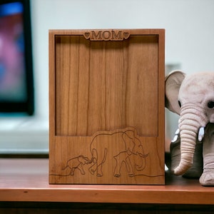 May include: A wooden picture frame with a cutout of three elephants and the word "MOM" with hearts on either side. The frame is sitting on a wooden table.