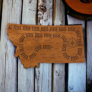 May include: A wooden game board shaped like the state of Montana with the names of cities and towns throughout the state. The board has a compass in the center and a grid of holes for playing a game.