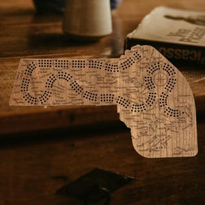 May include: Wooden cribbage board in the shape of a state, featuring drilled holes for pegs and engraved place names. The board is made of light-colored wood and has a compass rose design.