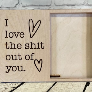 Puede incluir: Señal de madera con el texto "I love the shit out of you." con un corazón a cada lado del texto. La señal tiene un recorte para una foto.
