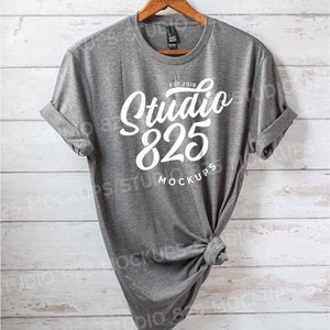 May include: A heather gray t-shirt with rolled-up sleeves hangs on a wooden hanger. The shirt features the white text "Studio 825 MOCKUPS" with "EST. 2019" above the main text. The shirt is knotted at the bottom.