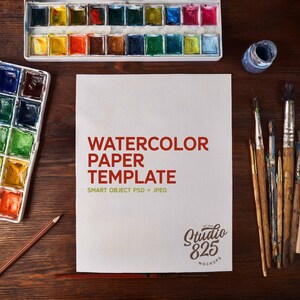May include: Overhead shot of watercolor art supplies on a wooden surface. Includes watercolor paper template with red text, paint brushes, pencils, and paint palettes filled with various colors. A small bottle of blue paint is also present.